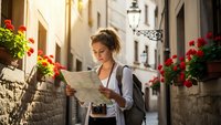 Young woman tourist with backpack reading map while exploring charming narrow cobblestone street with colorful flower boxes in historic old town district.