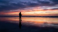 Silhouette of fisherman casting line at sunset on reflective beach with dramatic orange and purple sky clouds mirrored in wet sand creating peaceful moment.