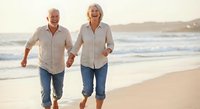 Happy senior couple running together on beach at sunset holding hands enjoying active retirement lifestyle and romantic vacation.