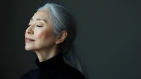 Serene mature Asian woman with silver gray hair and closed eyes wearing black turtleneck against dark background, expressing peace and mindfulness in profile view.