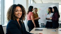 Professional mixed-race businesswoman with curly hair smiling in modern office meeting room with diverse female colleagues collaborating in background.