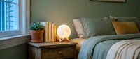 Cozy bedroom interior with warm lighting from bedside lamp, sage green pillows, books and plant on wooden nightstand creating peaceful atmosphere.