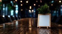Small white blank frame with decorative pine branches on wooden table against blurred bokeh lights background, creating cozy festive atmosphere.