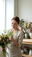 Young Caucasian female florist arranging fresh fern bouquet in bright workshop studio with natural light and plants on wooden counter for floral design.