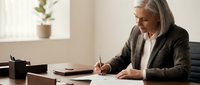 Mature woman with gray hair and light skin tone, wearing dark blazer and white shirt, writing on document at desk in modern office environment with plant in background.