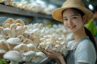 Young Asian female farmer in straw hat and apron harvesting fresh oyster mushrooms in modern indoor cultivation facility, showing sustainable agriculture.