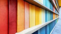 Colorful books arranged in rainbow order on white shelf creating vibrant perspective view. Spines form geometric pattern of red, orange, yellow and blue stripes.