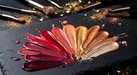 Professional makeup brushes with colorful lipstick swatches in red pink gold bronze tones on dark slate background for beauty cosmetics advertising.