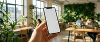 Hand holding smartphone with blank white screen in modern eco-friendly office workspace with green plants and natural lighting for mockup presentations.