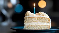 Slice of vanilla layer cake with white buttercream frosting and single lit blue birthday candle on dark plate against blurred bokeh background.