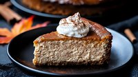 Creamy pumpkin spice cheesecake slice topped with whipped cream and cinnamon on dark plate, autumn leaves decoration, moody food photography.