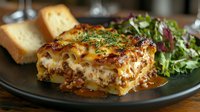 Traditional layered lasagna with meat sauce and melted cheese served on dark plate with fresh green salad and bread slices, close up view.
