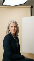 Mature woman with long gray hair wearing dark blazer seated in professional studio setting with soft lighting equipment and neutral background for corporate portraits.