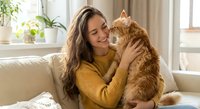 Young woman cuddling orange tabby cat at home, wearing yellow sweater on cozy sofa. Pet owner bonding with fluffy ginger cat in bright living room interior.