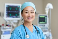 Confident Asian female medical professional in scrubs and surgical cap smiling warmly in hospital setting with monitoring equipment in background.