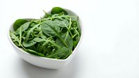 Fresh arugula and mixed green leaves in white heart-shaped bowl on white background, suitable for healthy eating, nutrition, and food styling projects.