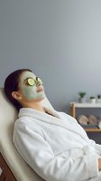 Young Asian woman relaxing in white bathrobe with green facial mask and cucumber slices on eyes during spa treatment at home wellness routine.