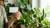 Person holding blank white card surrounded by lush green houseplants near window light, ideal for business cards, plant care tips, gardening services, or botanical product marketing.