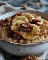 Creamy oatmeal topped with fresh banana slices, crunchy pecans, honey drizzle and chia seeds in white ceramic bowl. Healthy breakfast comfort food styling.
