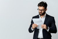 Bearded businessman in glasses and black suit reading document against light background. Professional male executive reviewing paperwork in corporate setting.