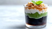 Layered dessert with green and black jelly, white cream and caramel sauce topped with nuts and mint in glass cup for gourmet presentation and culinary photography.