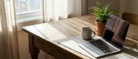 Wooden desk with laptop, notebook, and potted plant near window with natural sunlight for home office or workspace design projects.