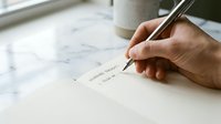 Person writing with pen on white paper at marble desk, close-up of hand signing document or letter, workspace with coffee cup, business correspondence and administrative tasks.