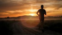 Male runner jogging on rural road at sunrise with golden sky and misty landscape. Silhouetted athlete in motion during early morning workout outdoors.