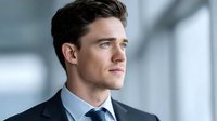 Professional young Caucasian businessman in dark suit and tie looking confidently ahead in modern office environment with natural lighting.