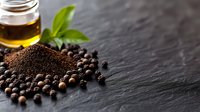 Ground and whole black peppercorns with fresh basil leaf and olive oil on dark stone background, macro food photography for cooking and seasoning.