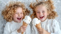 Joyful children with curly blonde hair lying on gray carpet, wearing white sweaters and laughing while holding vanilla ice cream cones, expressing pure happiness and fun.
