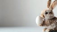 Adorable baby bunny holding white egg against neutral gray background, perfect for Easter holiday designs and spring season promotions.