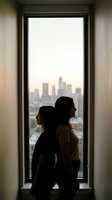 Two women silhouetted against large window overlooking city skyline at sunset, intimate moment of connection and contemplation in modern apartment.