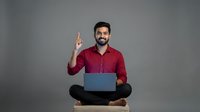 Smiling bearded man in red shirt pointing up with laptop computer sitting cross-legged on gray background for business presentations and marketing materials.