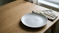 Empty white plate with folded napkin on wooden table near window, minimalist dining setup for restaurant, home, or food service design projects.