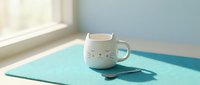 White ceramic cat-shaped mug with whiskers and ears on turquoise placemat by sunny window. Cute kitty coffee cup for morning beverage, home decor and kitchen accessories.