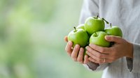 Unrecognizable person holding fresh green apples in hands against blurred natural background, promoting healthy eating and organic fruit harvesting.