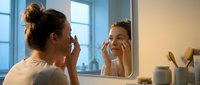 Woman applying skincare moisturizer to face while looking in bathroom mirror during morning routine, wellness and beauty care at home.