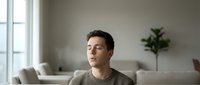 Young man practicing mindfulness meditation at home, sitting peacefully with eyes closed in modern living room with natural light from window.