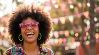 Happy African American woman with curly afro hair wearing pink glasses laughing joyfully outdoors with colorful bokeh lights background for lifestyle content.