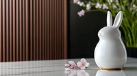 White ceramic bunny figurine with gold base on wooden table with delicate pink cherry blossoms, against dark striped wall background. Modern minimalist spring decor setting.