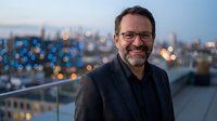 Smiling middle-aged Caucasian businessman with glasses and beard on modern rooftop terrace at dusk with blurred city lights background for corporate success.