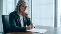 Mature businesswoman with gray hair and glasses reviewing documents at office desk, professional executive analyzing paperwork in corporate meeting room.