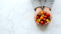 Overhead view of hands holding fresh cherry tomatoes in various red and yellow hues on white marble surface for cooking and recipe content.