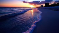 Stunning sunset over ocean waves washing onto pristine white sand beach with coastal buildings silhouetted against dramatic purple and orange sky.