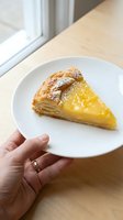 Hand holding white plate with golden custard tart slice on wooden table near window for breakfast dessert presentation.