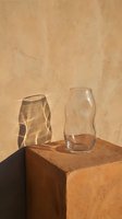 Two empty glass vessels sit on a terracotta cube against a warm beige textured wall, casting geometric shadows in natural sunlight for minimalist still life compositions.