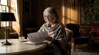Senior Caucasian woman with glasses reading documents at wooden desk in elegant home office with warm lighting and classic decor for business content.