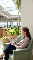 Young professional woman working on tablet in modern office space with plants and natural light from skylights for remote work and productivity.