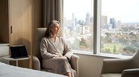 Mature Caucasian businesswoman in beige suit sitting by hotel window overlooking city skyline, contemplating during business travel or corporate meeting.